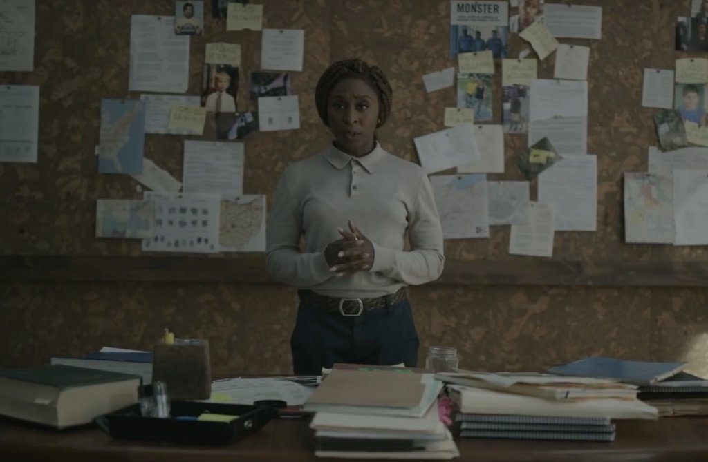 Holly presents her findings, evidence pinned to the wall behind her