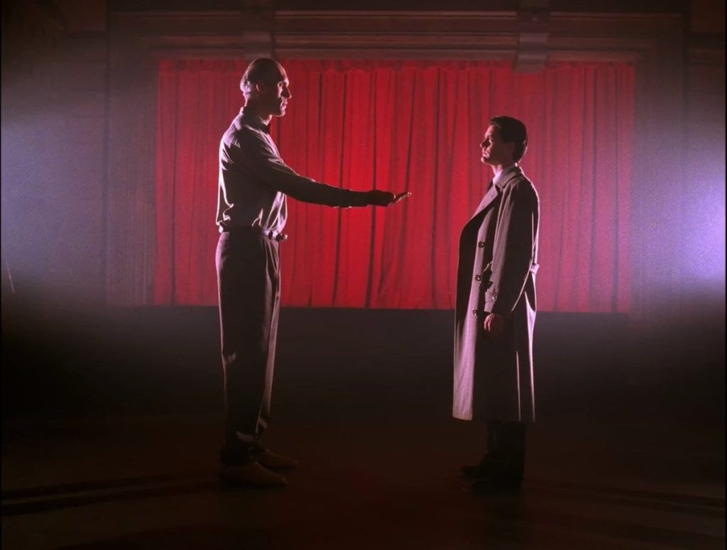 Dale Cooper and The Giant stand facing each other in front of a red curtain and The Giant extends his hand to Cooper.