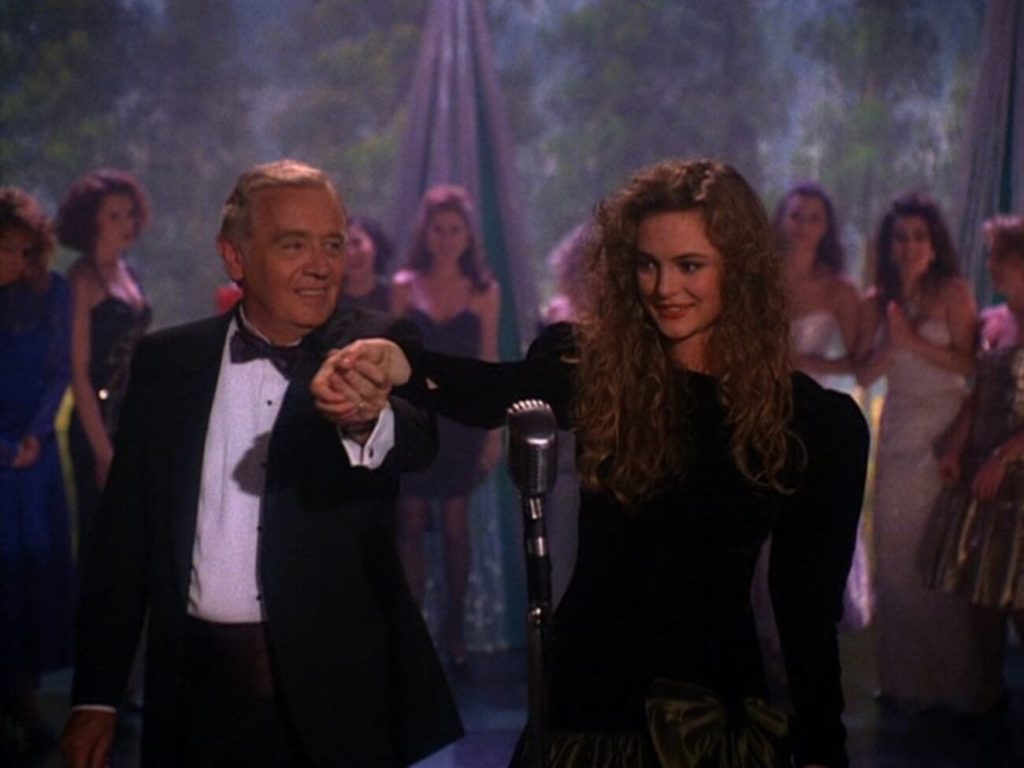 Annie celebrates winning the Miss Twin Peaks pageant on stage, in front of a microphone, her hand raised with help from Milford